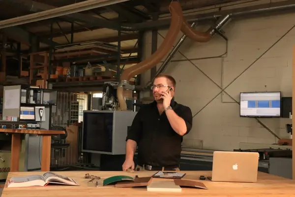 Ein Mann in einer modernen Werkstatt telefoniert am Schreibtisch mit Laptop und Plänen.