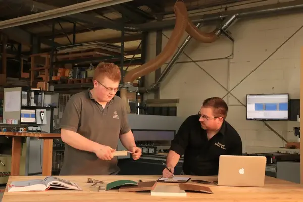 Zwei Männer besprechen Holzarbeiten mit technischen Unterlagen in einer modernen Werkstatt.