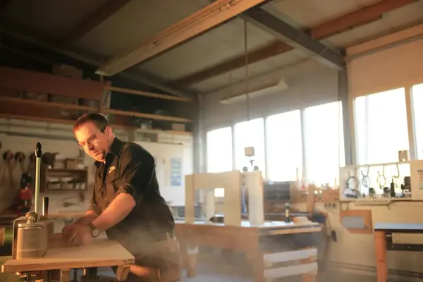 Ein Handwerker schleift Holz an einer Werkbank in einer sonnendurchfluteten Werkstatt.
